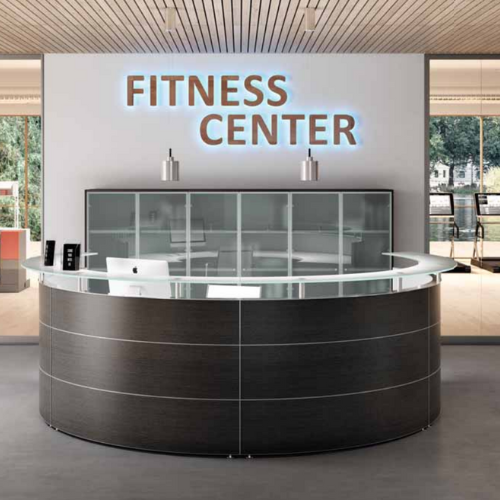 Round reception desk in a dark wood, matching cabinets behind. The Reception has lighting behind which says 'fitness center'